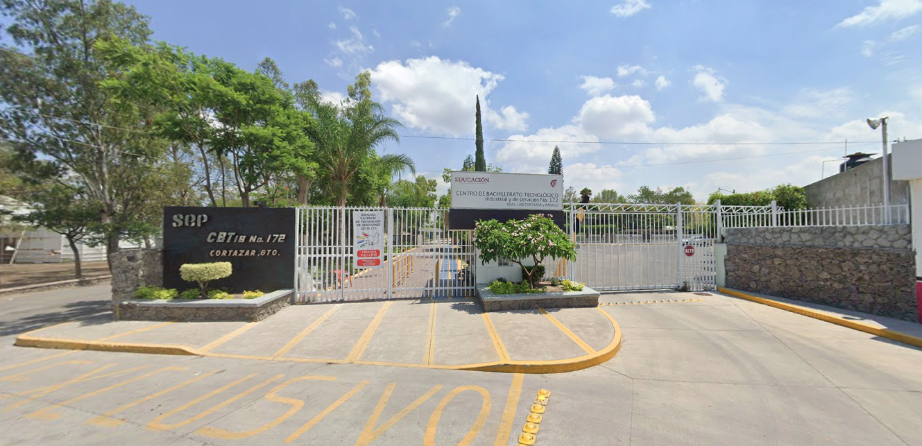Fotografía en la que se muestra un muro café y sobre él está escrito el nombre del plantel. Al lado derecho del muro hay una reja blanca por la cual se puede ingresar caminando. Al terminar la rej<a hay una caseta y otra reja blanca que da acceso al estacionamiento.